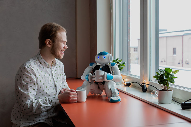 En person sitter vid ett bord och dricker kaffe samtidigt som den ser ut att ha en konversation med en robot.