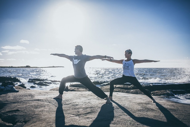 Två personer står på en strand med havet i bakgrunden och gör yoga.